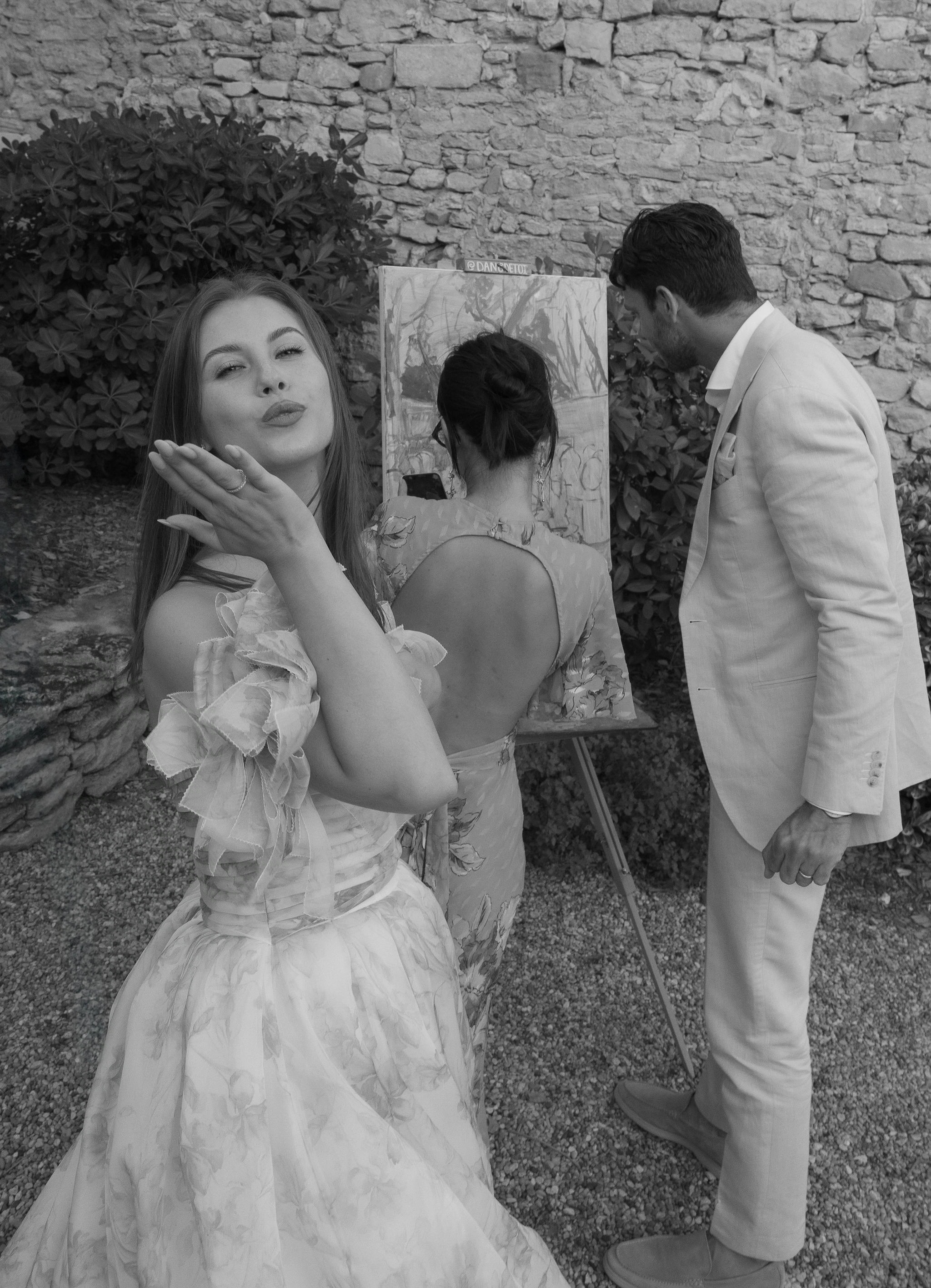 Bride blowing a kiss at the camera while the groom watches their wedding painting take shape on the easel. © Danielle Villarreal