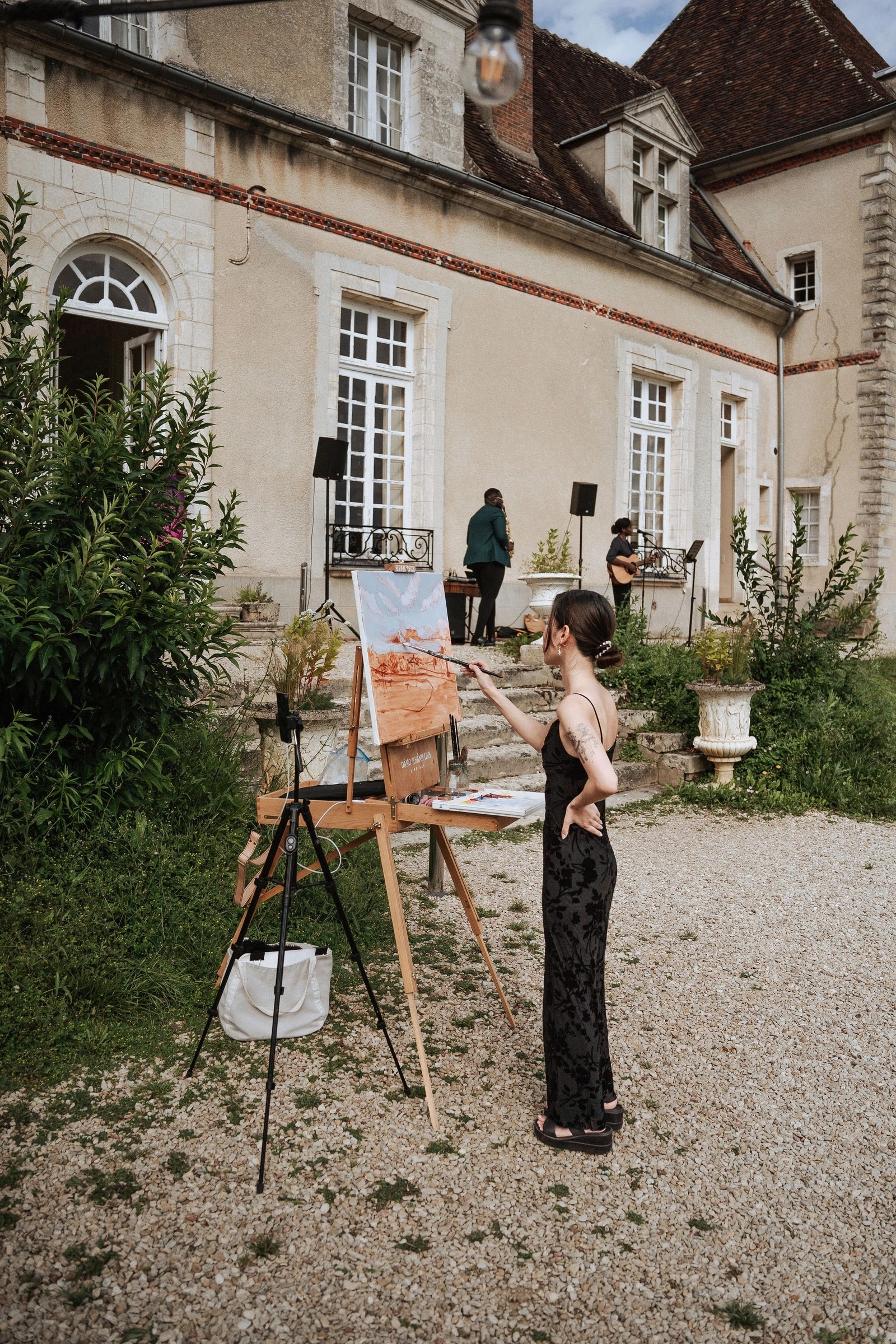 Hochzeitsmalerin Linh Dang arbeitet an einer Leinwand, während eine Sängerin und un Saxophonist im Hintergrund des Château du Feÿ auftreten. Foto: Kimmak Design.