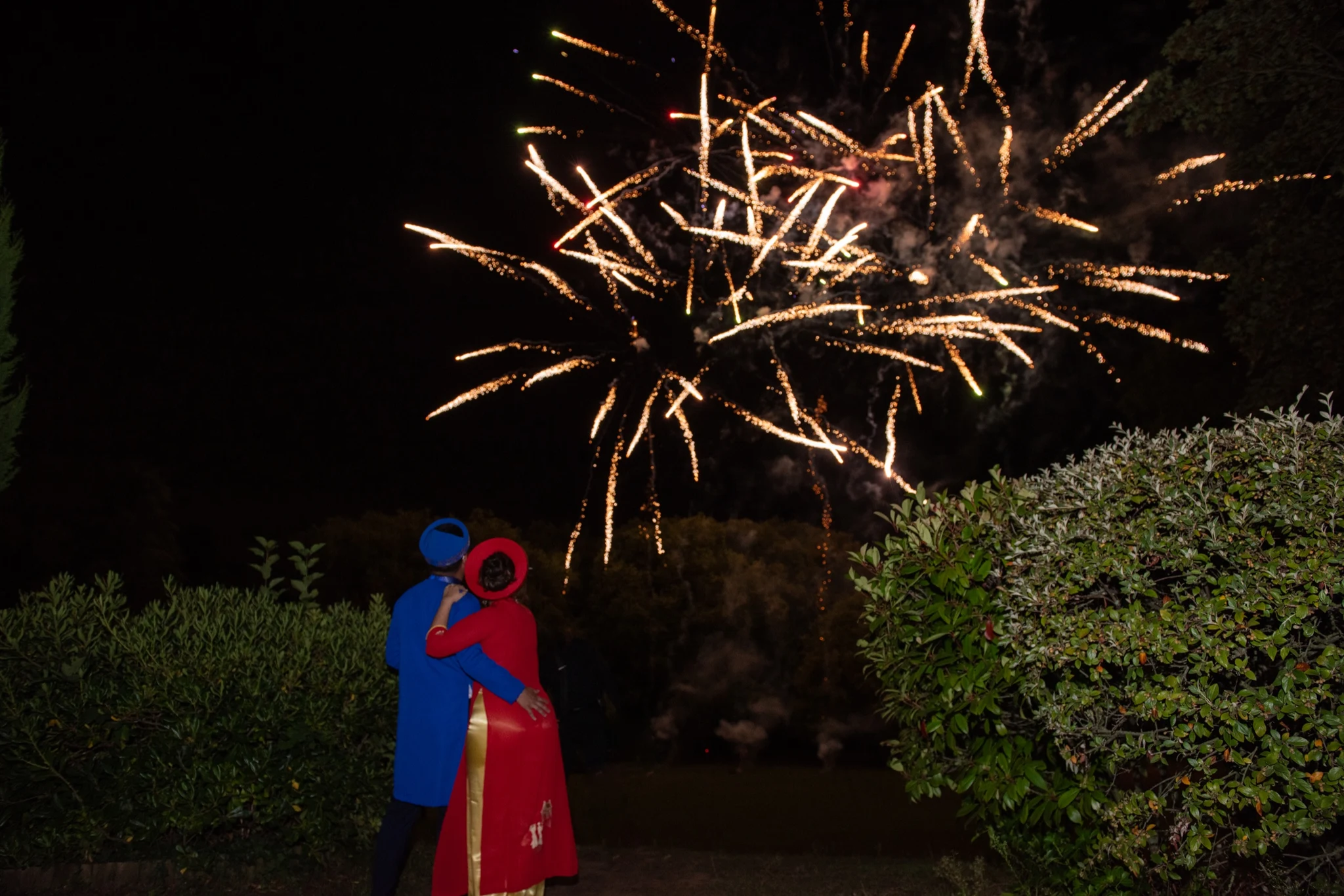 Bride and groom in red and blue ao dai hugging under fireworks at Château de la Tour. Photography by GC Photographie.