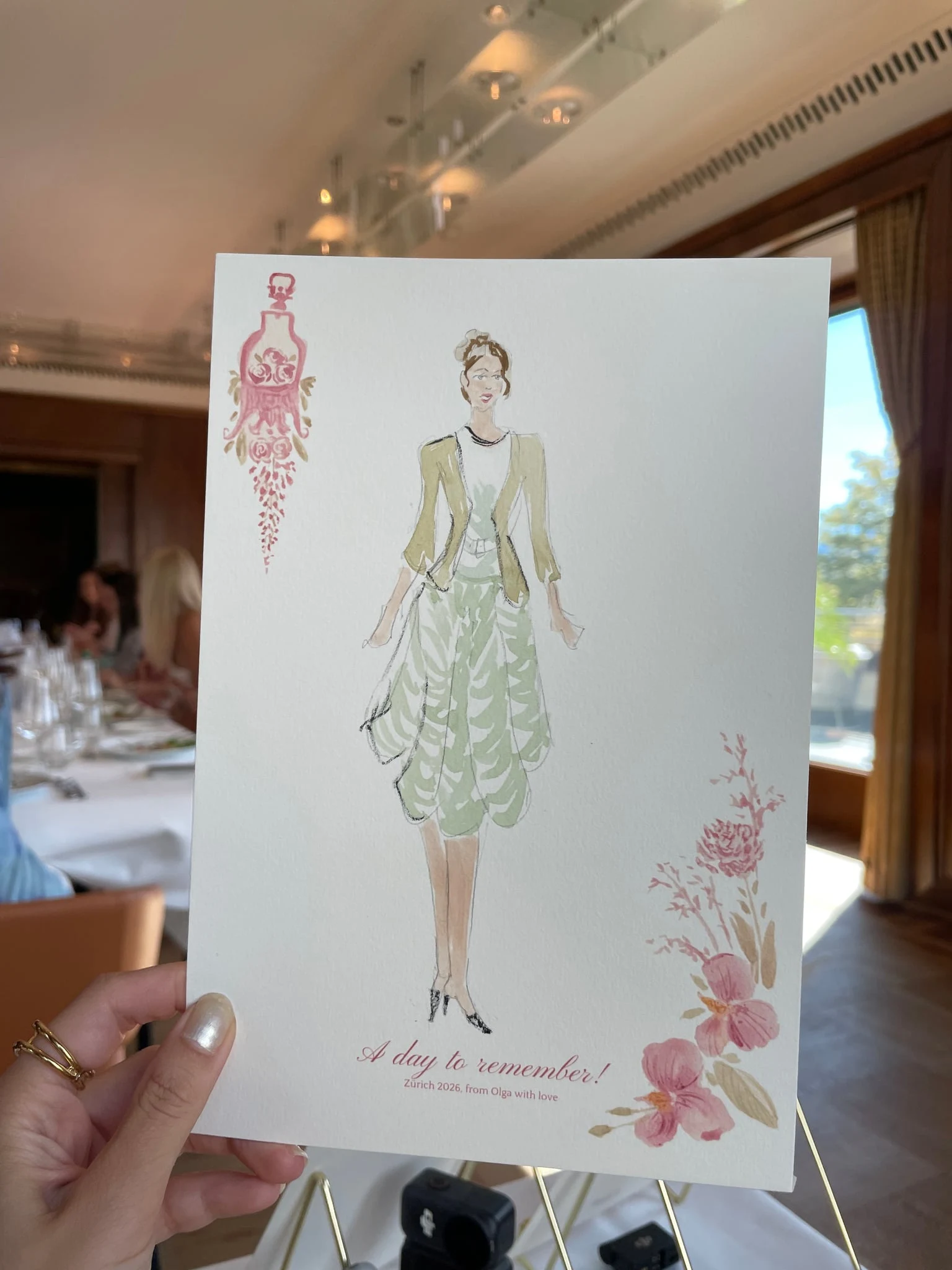 Live watercolor illustration of a guest in a sage dress and olive jacket at a birthday party at AMERON Zürich Bellerive au Lac.